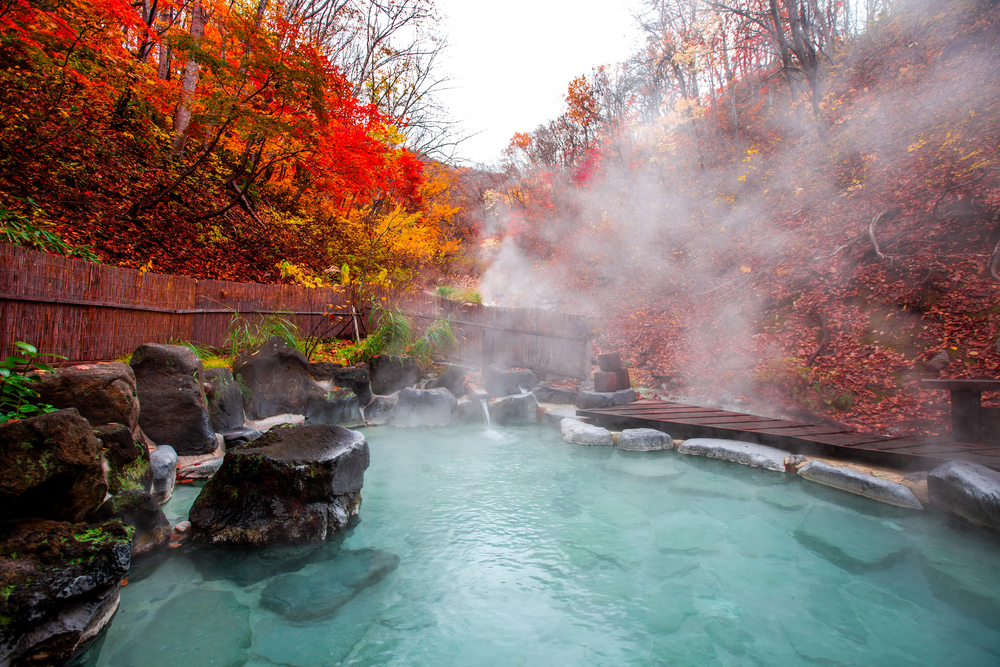Steaming hot spring surrounded by rocks and autumn trees with red and yellow leaves, capturing the serene beauty of traveling in Japan.