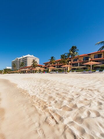 Sandy beach with turquoise water, palm trees, and beachfront hotels under a clear blue sky—experience the ultimate Grand Cayman luxury travel escape.