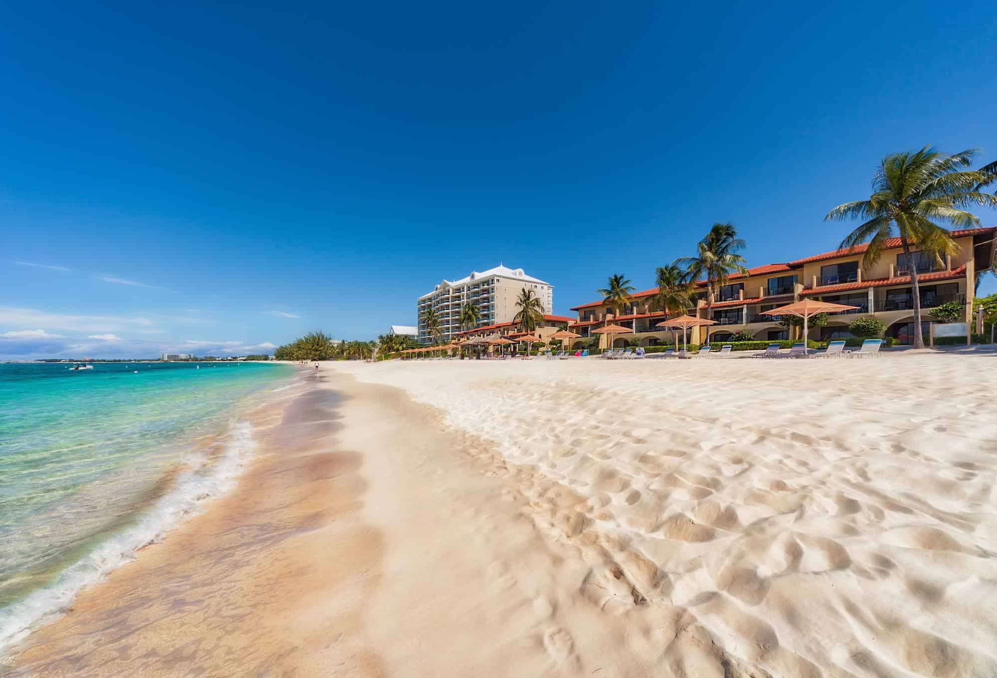 Sandy beach with turquoise water, palm trees, and beachfront hotels under a clear blue sky&mdash;experience the ultimate Grand Cayman luxury travel escape.