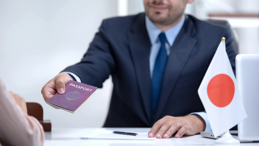 Man in suit hands a passport to someone; Japanese flag is on the desk in front of him, suggesting preparations for traveling in Japan.
