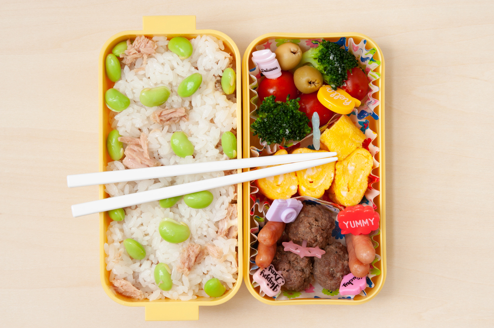Yellow bento box with rice, edamame, tuna, veggies, eggs, meatballs, sausages, and chopsticks on top—perfect for enjoying a delicious meal while traveling in Japan.