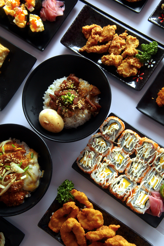 Assorted sushi, rice bowls, and fried chicken dishes arranged on black plates evoke the vibrant culinary experiences you’ll find while traveling in Japan, beautifully displayed on a white surface.