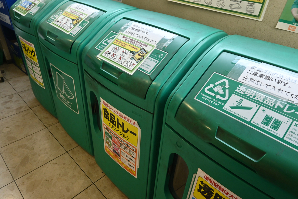 Four green recycling bins with labels and signs in Japanese, lined up against a tiled wall indoors—a common sight when traveling in Japan.