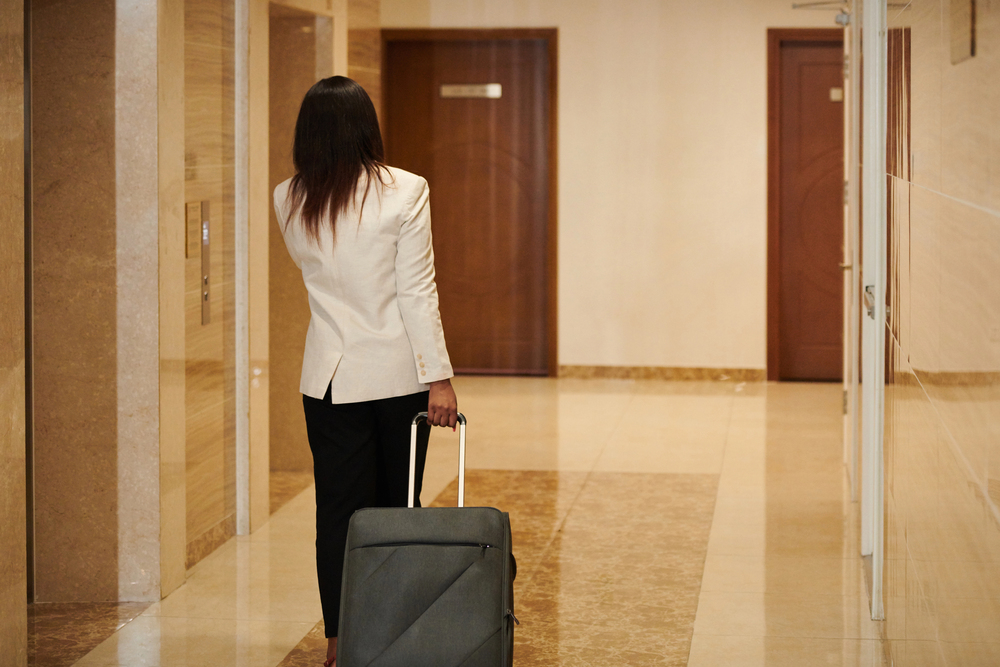 A woman with a suitcase stands in a hotel hallway facing elevator doors, dressed in a white blazer, as she prepares for another day traveling in Japan.