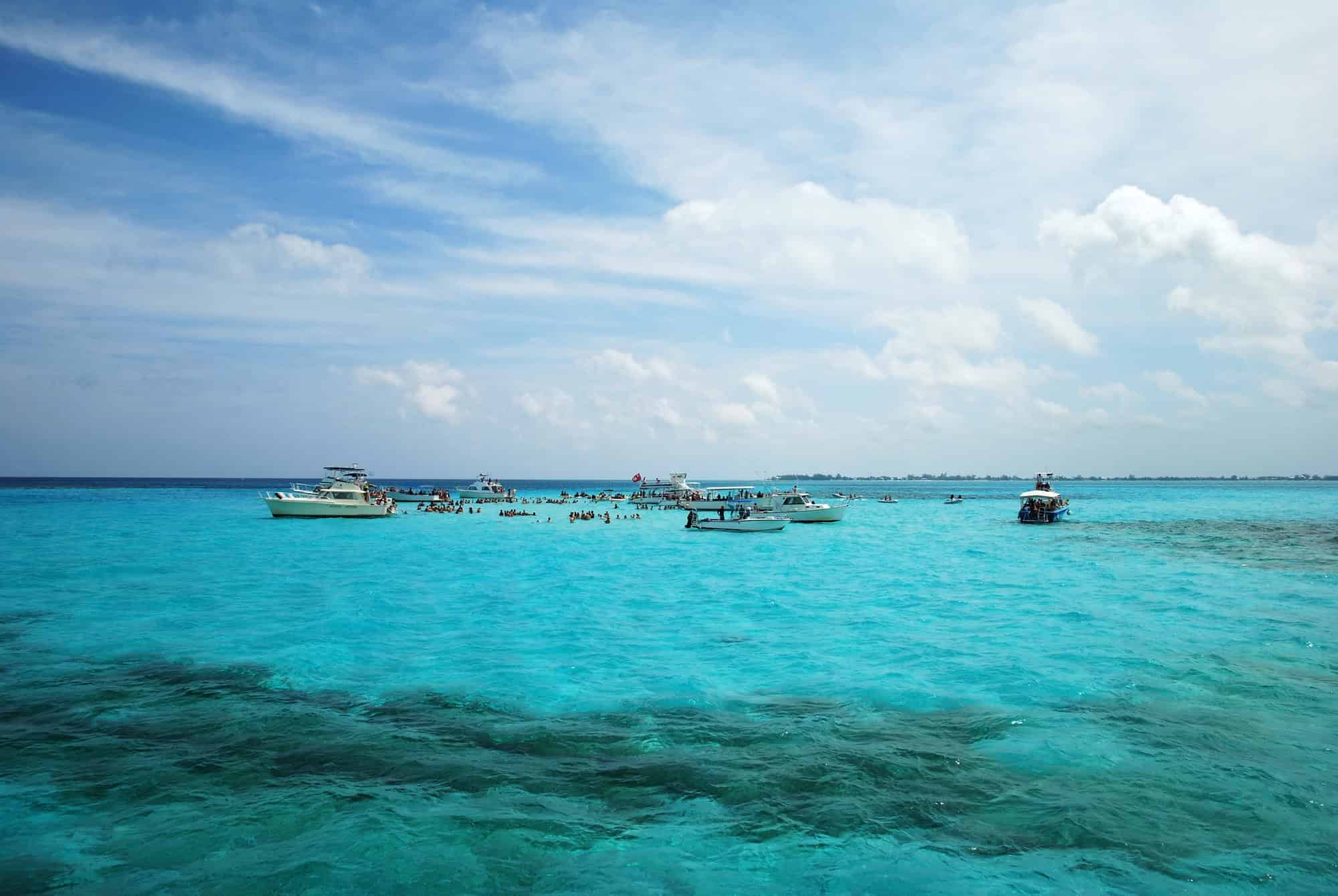 Several boats float on bright turquoise water under a partly cloudy sky, with people swimming nearby&mdash;offering an idyllic scene that captures the essence of Grand Cayman luxury travel.