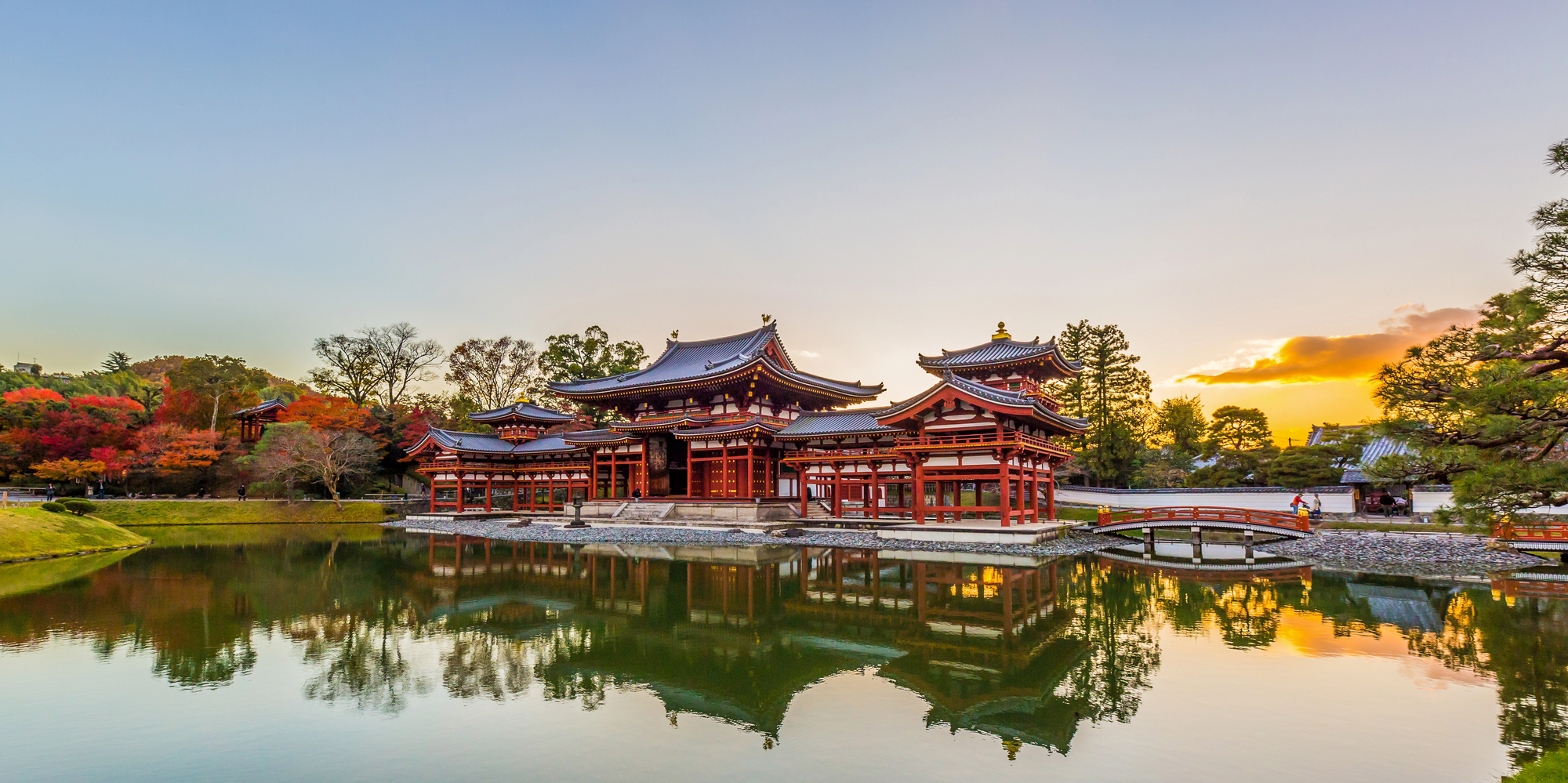 Traditional Japanese temple with curved roofs reflected in a pond at sunset, surrounded by trees and a bridge—a serene highlight for anyone traveling in Japan.