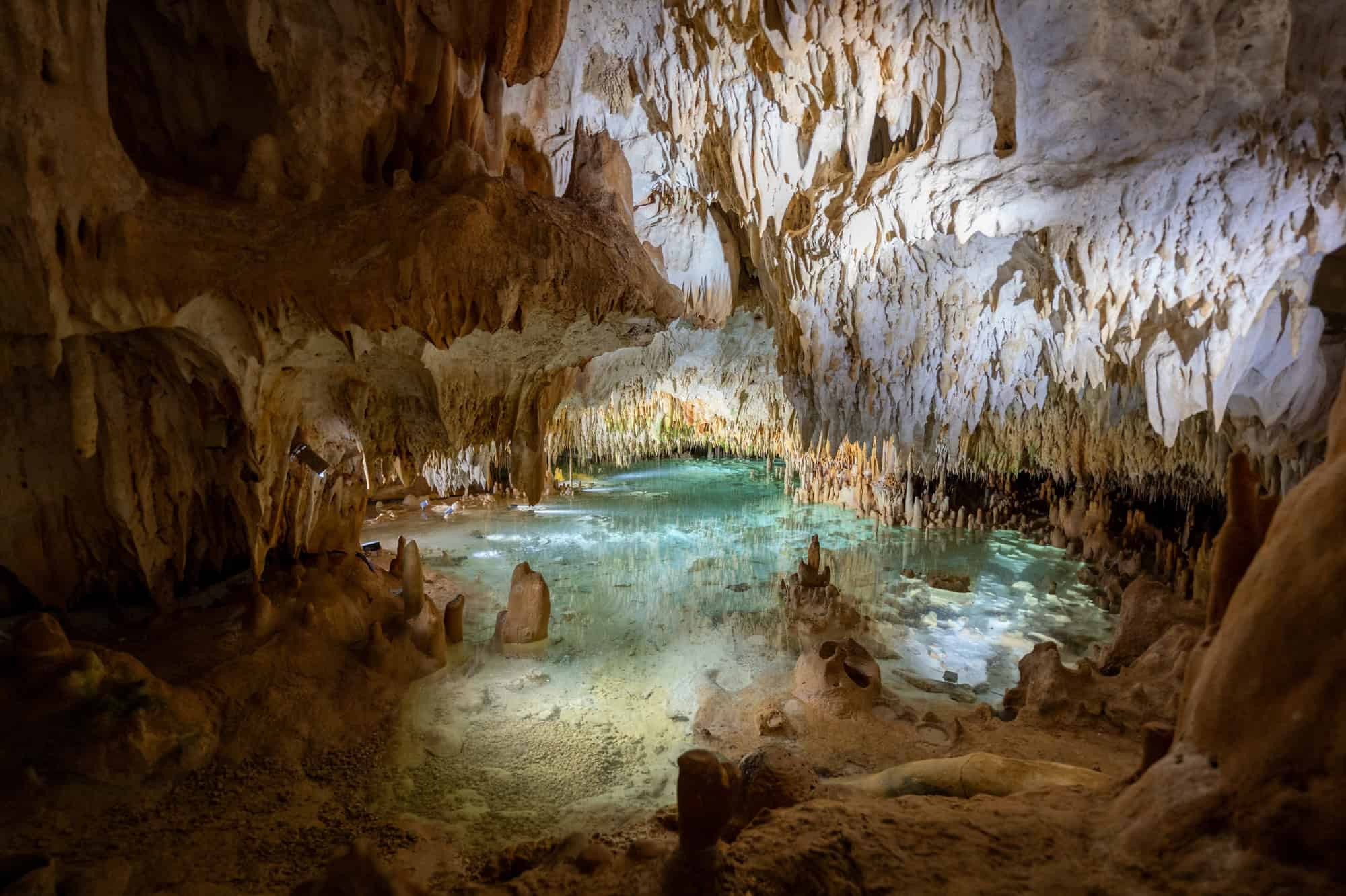 A cave with stalactites, stalagmites, and a pool of clear, turquoise water lit by soft light evokes the serene allure of Grand Cayman luxury travel.