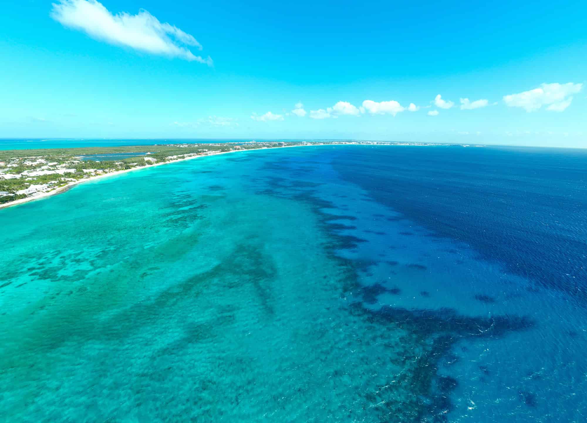 Aerial view of a turquoise ocean meeting the shoreline under a bright blue sky with a few scattered clouds, capturing the essence of Grand Cayman luxury travel.