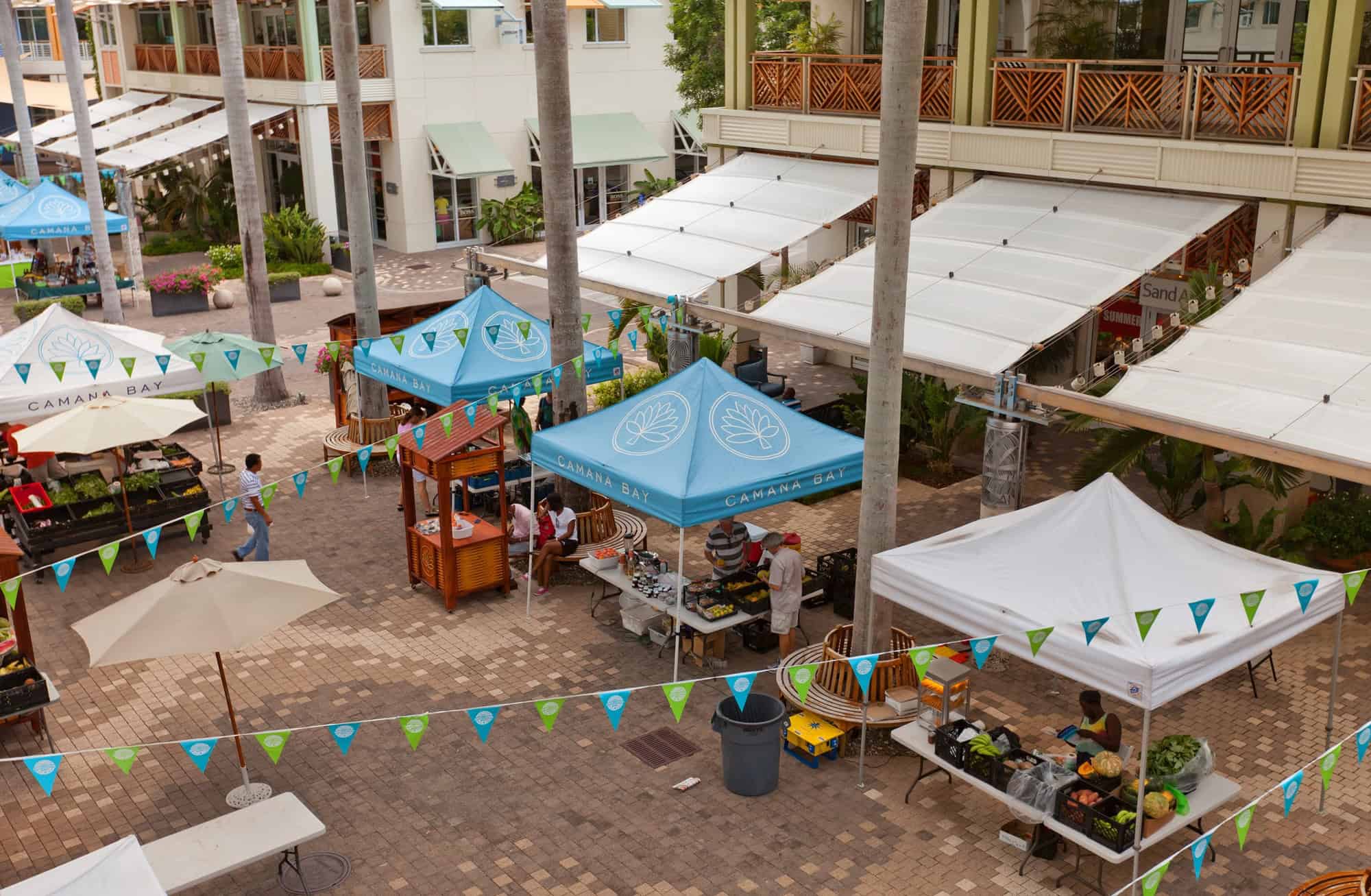 Outdoor farmers market with white and blue tents, vendors, and produce stands lining a tiled plaza&mdash;a charming highlight for those seeking authentic local experiences during Grand Cayman luxury travel.
