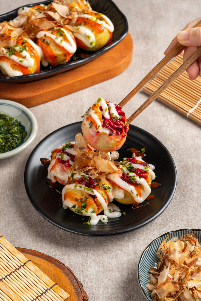 Chopsticks holding takoyaki topped with sauces, bonito flakes, and herbs over a black plate—a must-try experience when traveling in Japan.