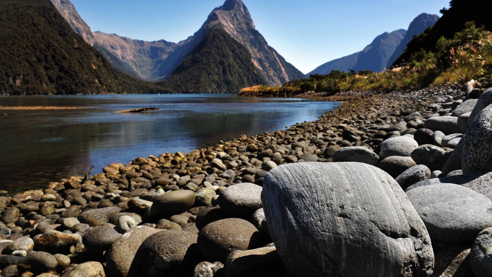 Rocky shoreline with smooth stones, calm water, and tall mountains under a clear blue sky.