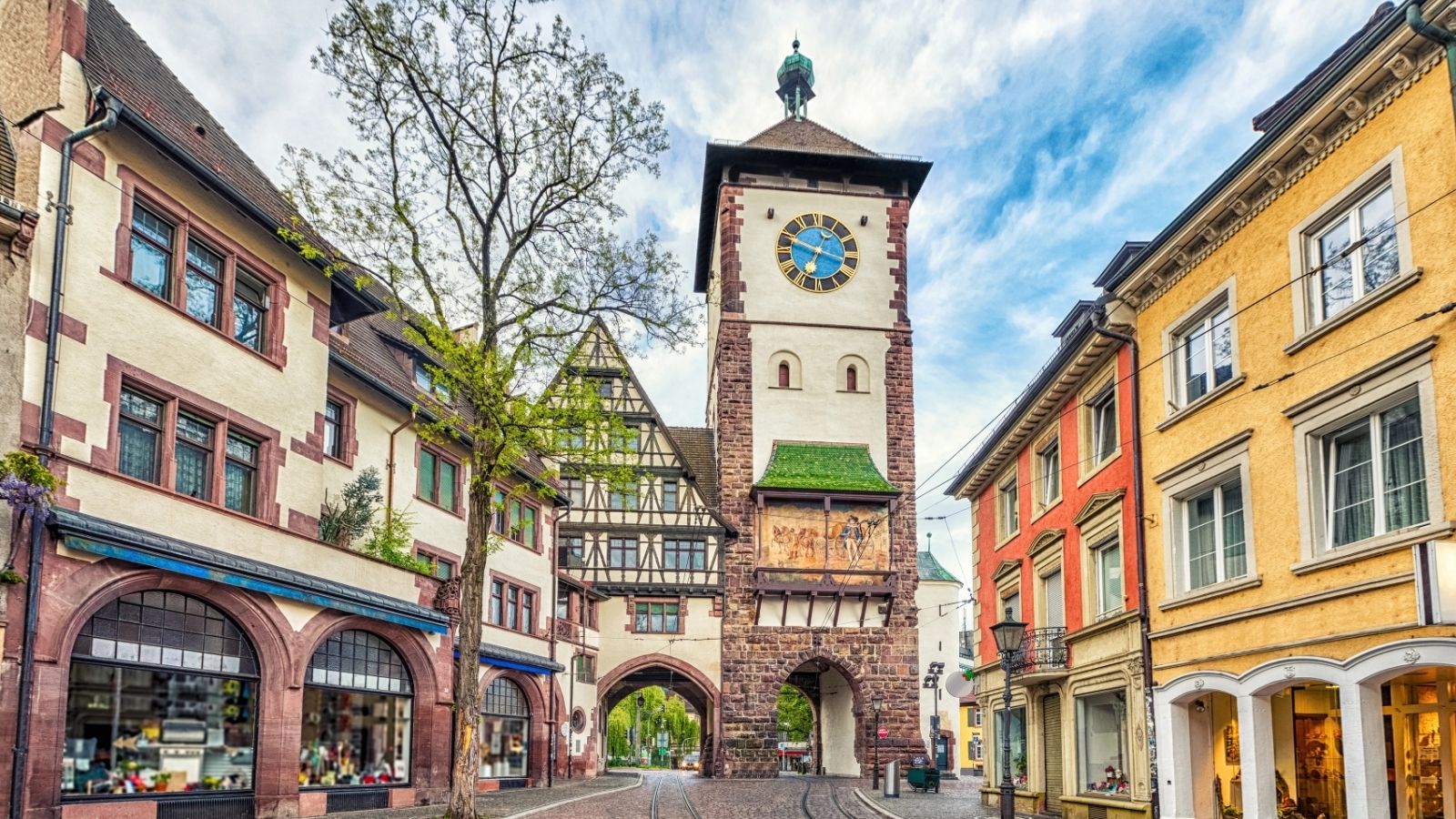 Colorful historic buildings and a tall clock tower frame a cobblestone street under a partly cloudy sky.