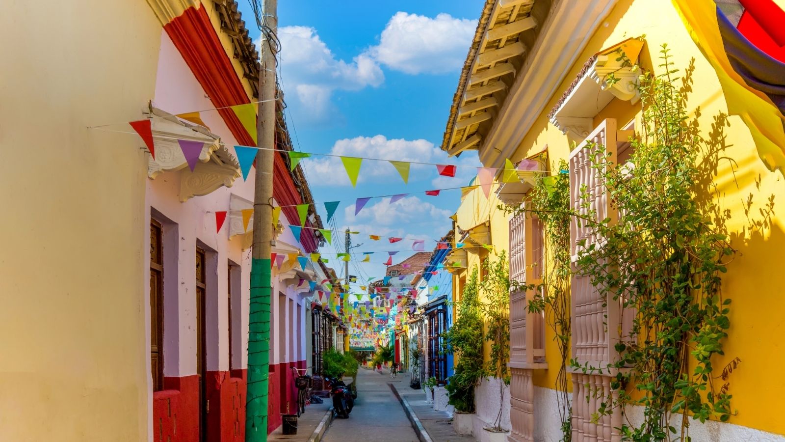 Colorful houses and festive pennant banners line a narrow street under a bright blue sky with fluffy clouds.