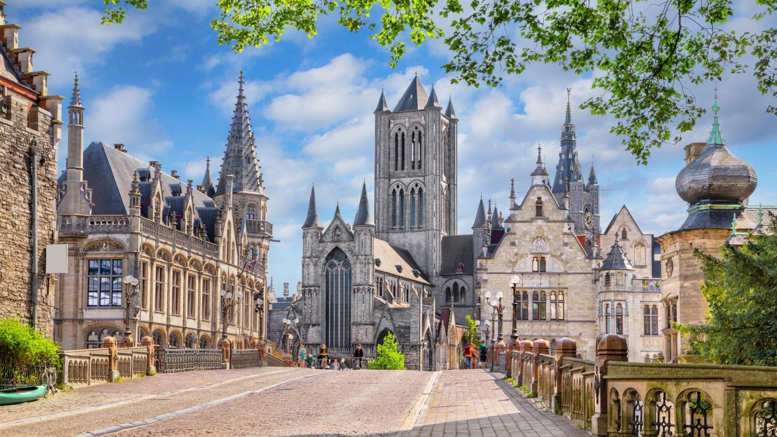 Medieval buildings and church towers line a cobblestone street under a blue sky with scattered clouds.