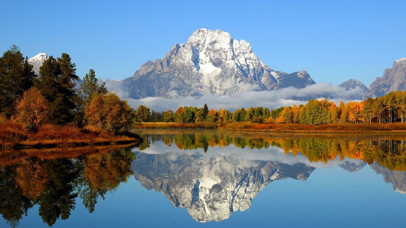 Snowy mountain reflected in a calm lake, surrounded by autumn trees and a clear blue sky.