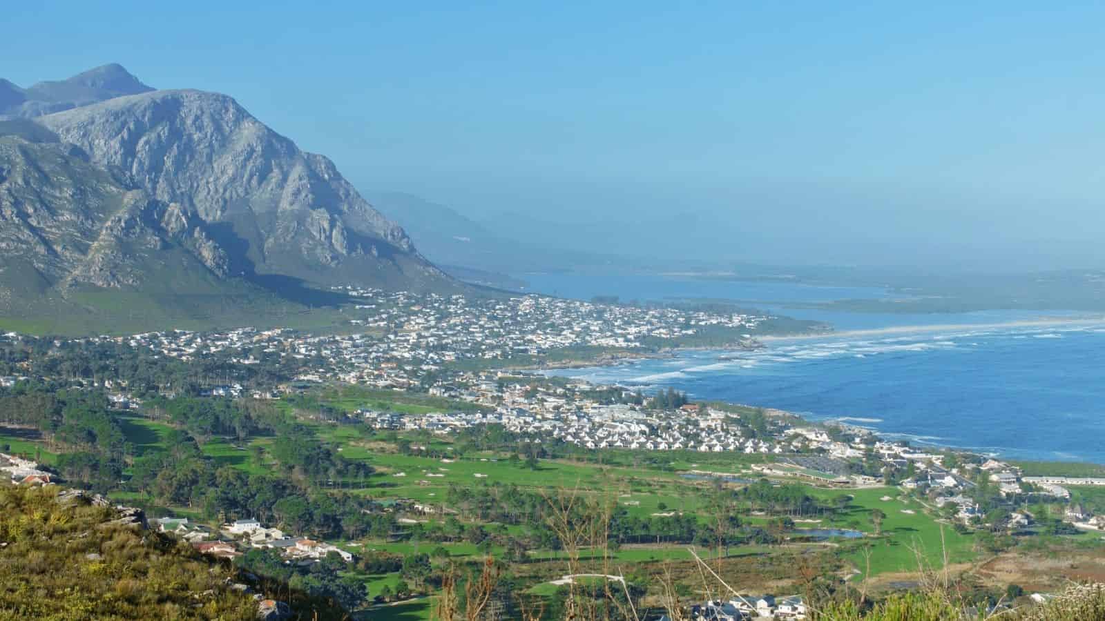 Coastal town with mountains, green fields, and blue sea under a clear sky.