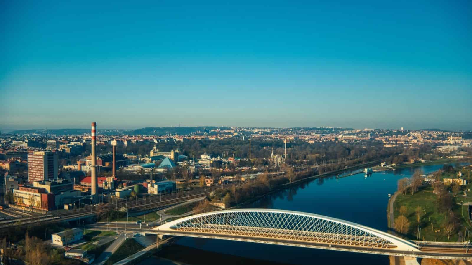 A modern white bridge spans a calm river with a city and smokestack in the background under a clear sky.