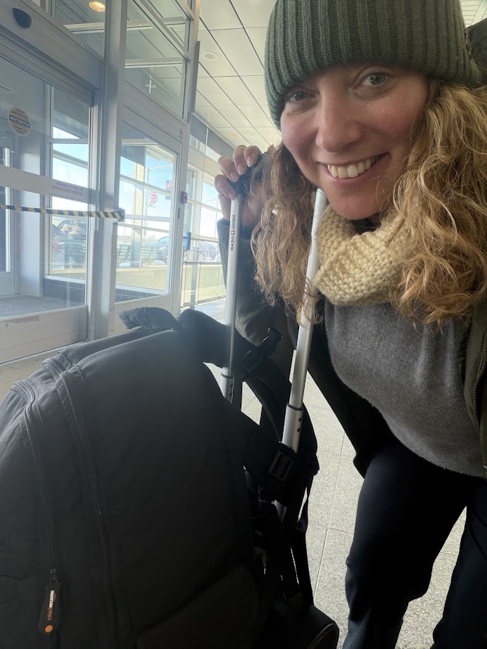 Smiling woman with a knit scarf and beanie, holding walking poles next to luggage in an airport, ready to embark on her Churchill northern lights tour.