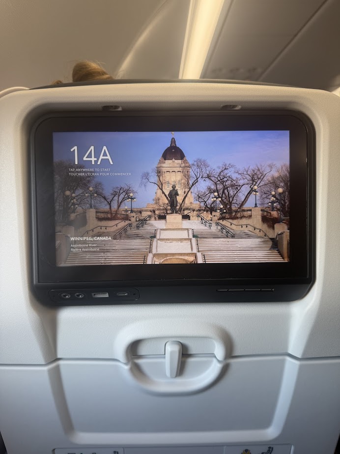 Airplane seat-back screen showing 14A with an image of a building and statue in Winnipeg, Canada, inviting travelers to embark on a Churchill northern lights tour.