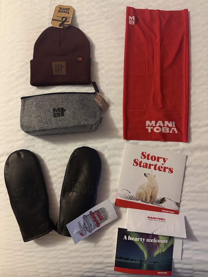 Various Manitoba-themed items on a bed, including mittens, beanie, pouch, scarf, booklets, and a postcard&mdash;perfect keepsakes to remember your Churchill northern lights tour.