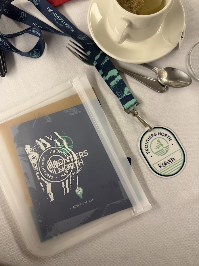Frontiers North lanyard, badge labeled "Kevin," adventure map&mdash;perhaps plotting a Churchill northern lights tour&mdash;alongside a tea cup and utensils on a white tablecloth.