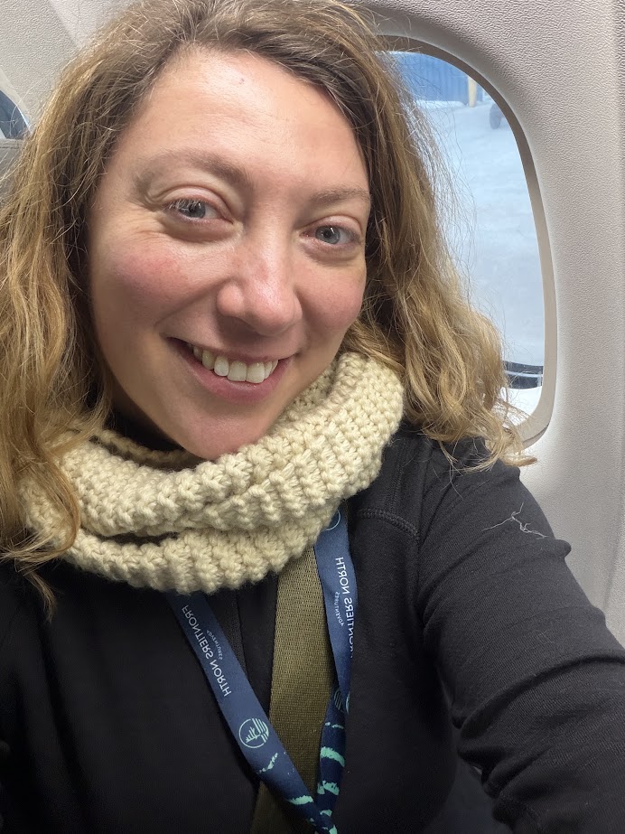 Woman with wavy hair smiles, wearing a knit scarf and lanyard, seated by an airplane window&mdash;ready to embark on a Churchill Northern Lights tour.