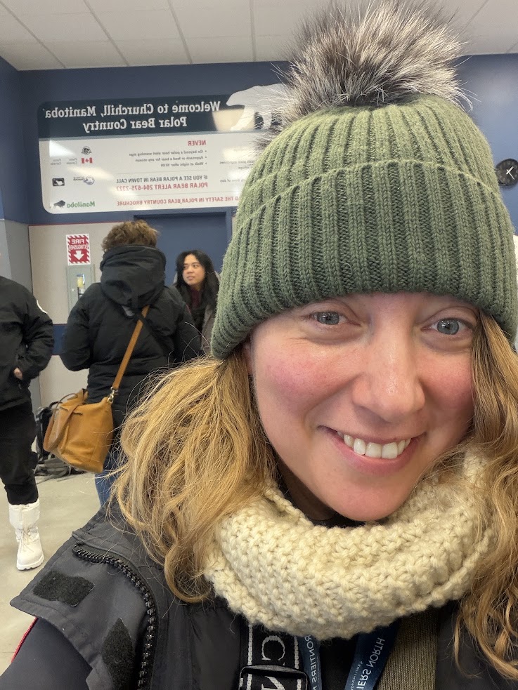 Woman in winter clothing smiles indoors after an exciting Churchill Northern Lights tour; people and a sign are visible in the background.