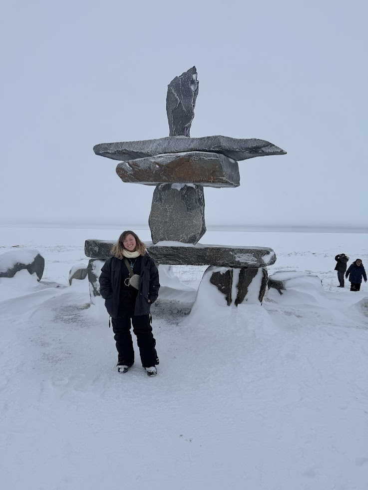 Person stands in snow by a large inuksuk; snowy landscape and a few people enjoying the view in the background, capturing the magic of a Churchill northern lights tour.