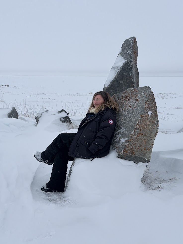 Person in a black winter coat sits on snowy rocks in a frozen, foggy landscape, capturing the serene beauty often witnessed during a Churchill northern lights tour.