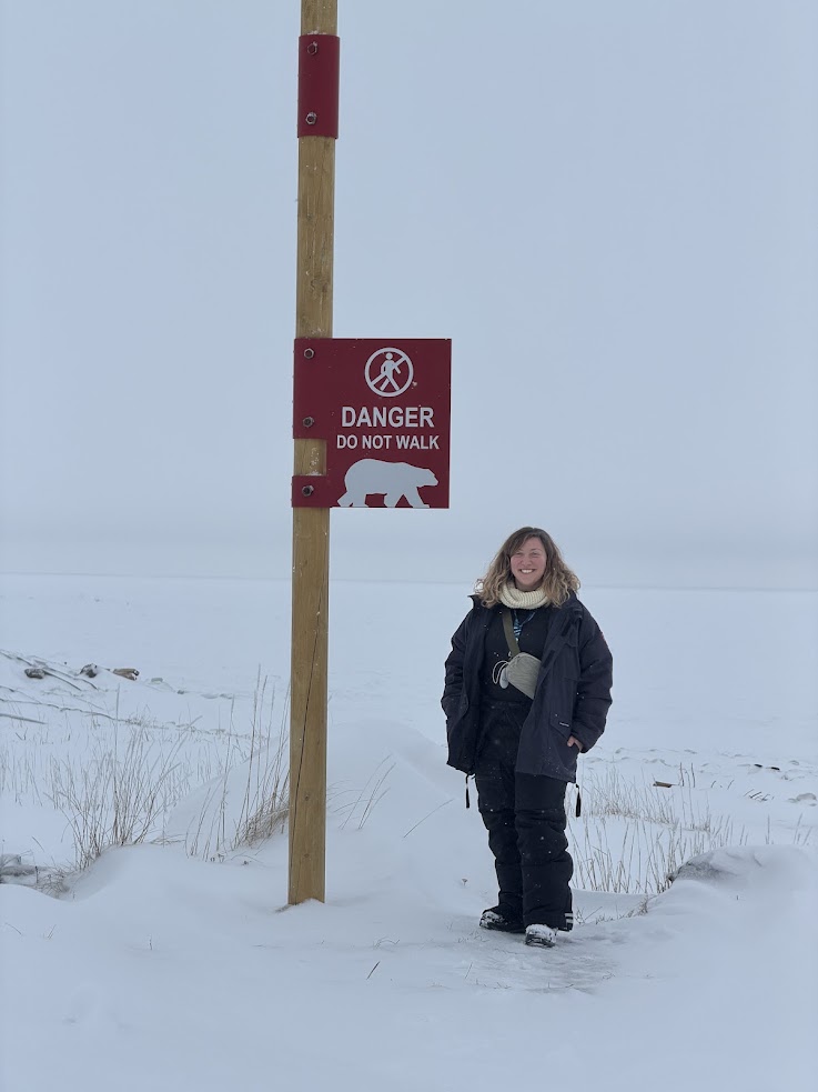 Person stands beside a "Danger: Do Not Walk" sign on a snowy landscape with overcast sky, capturing the stark beauty often experienced on a Churchill northern lights tour.