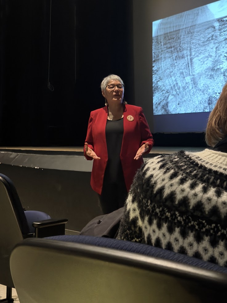 A woman in a red blazer speaks in front of an audience, with a large screen behind her displaying an image from the Churchill Northern Lights tour.