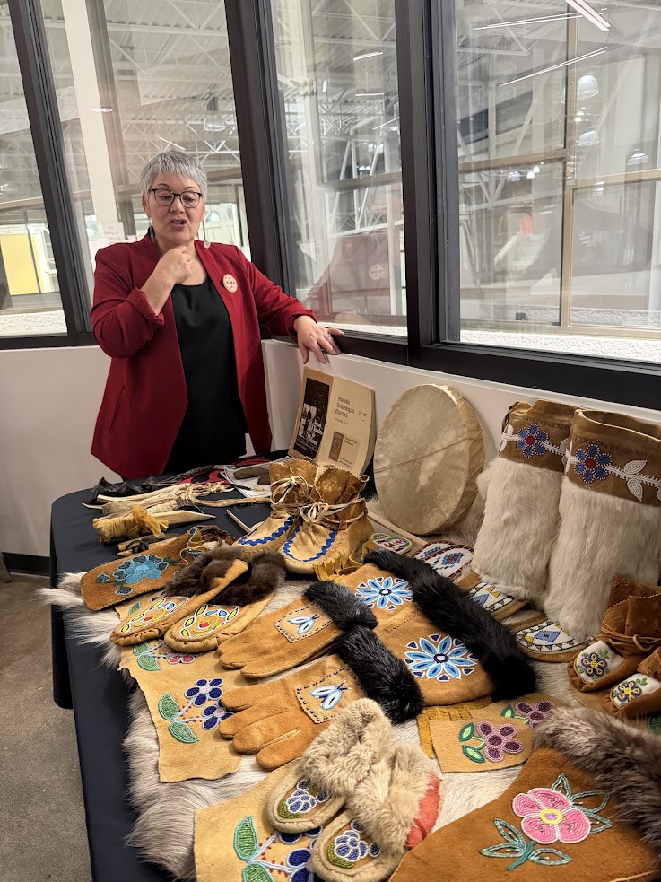 A woman stands by a table displaying colorful, beaded Indigenous crafts, gloves, and moccasins&mdash;beautiful souvenirs to remember your Churchill northern lights tour.