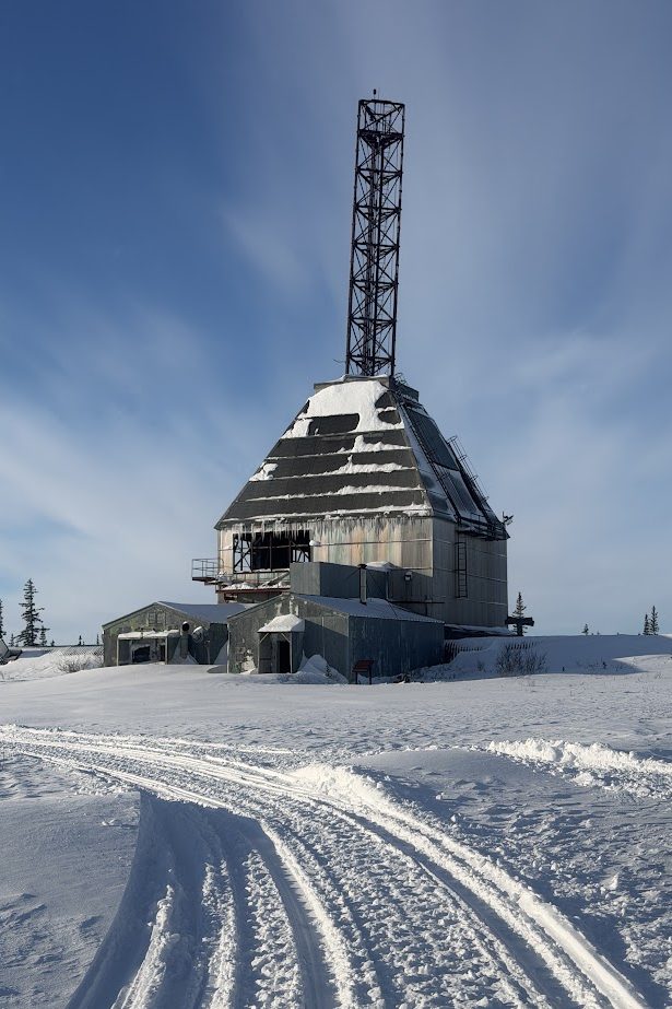 A large, snow-covered industrial building with a tall tower stands in a wintry landscape under blue sky, reminiscent of the stark beauty seen on a Churchill northern lights tour.