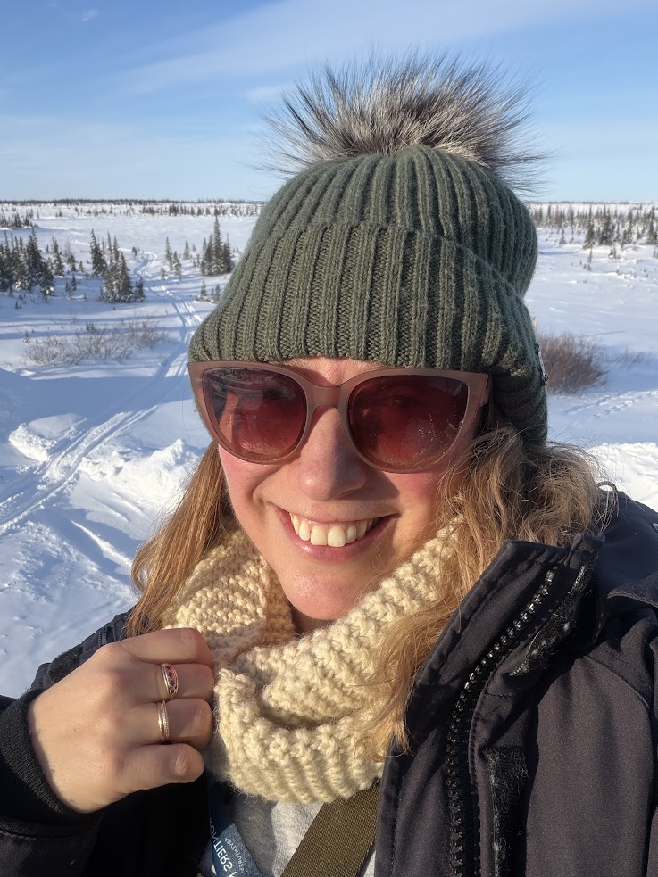 Smiling person in winter clothes and sunglasses, standing outside in a snowy, sunny landscape, perhaps ready to embark on a Churchill Northern Lights tour.