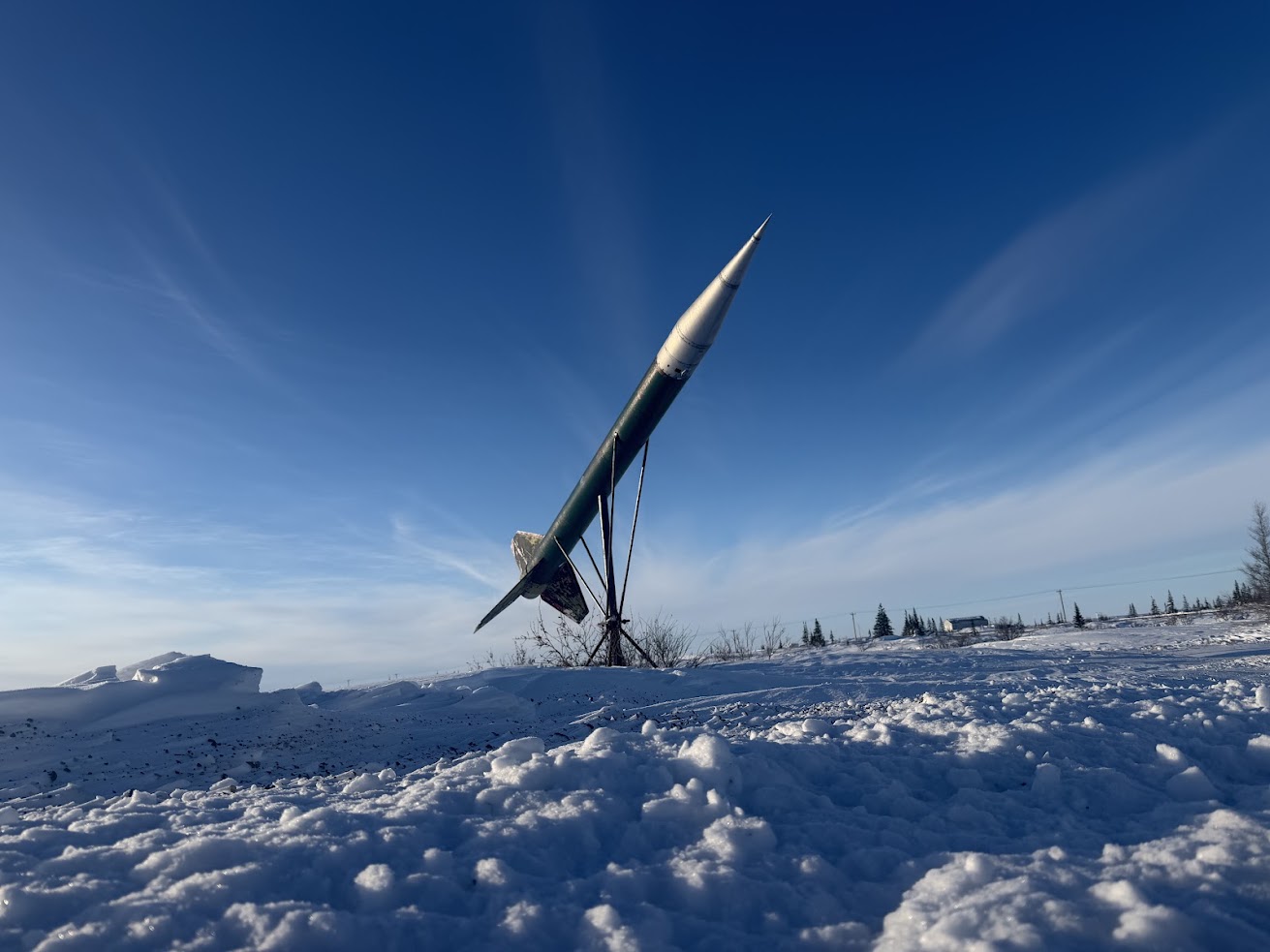 A tall rocket sculpture stands pointed skyward on a snowy landscape under a clear blue sky, reminiscent of the breathtaking views you might see on a Churchill northern lights tour.