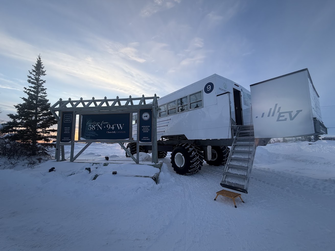 Large white off-road vehicle with stairs parked in snowy landscape, near a sign and a tree at sunset, ready for a Churchill Northern Lights tour.