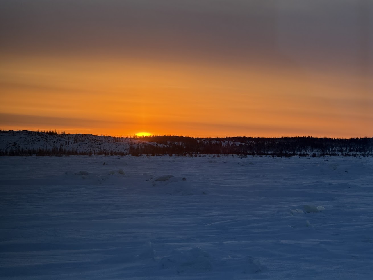 Sunset over a snowy, frozen landscape with an orange sky and a distant tree line, reminiscent of vistas seen on a Churchill northern lights tour.