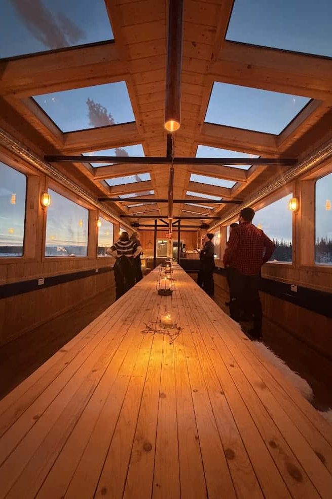 People standing in a cozy, wooden room with skylights and a long table lit by warm orange lights, reminiscent of gatherings after an adventurous Churchill Northern Lights tour.