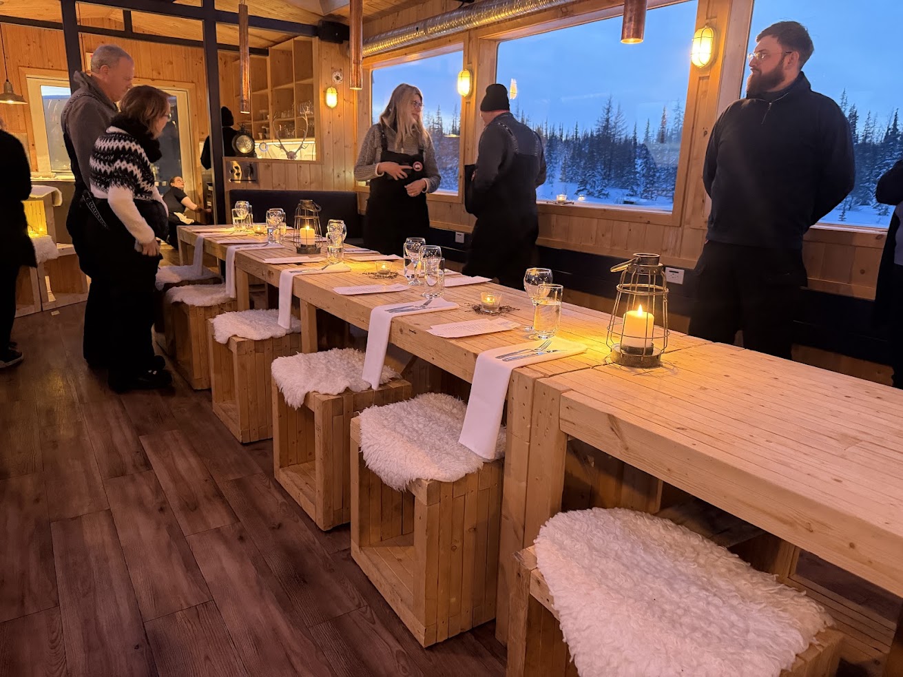 Wooden dining table set for dinner in a cozy cabin, with people standing and talking inside&mdash;much like the warmth you feel after an evening on a Churchill northern lights tour.
