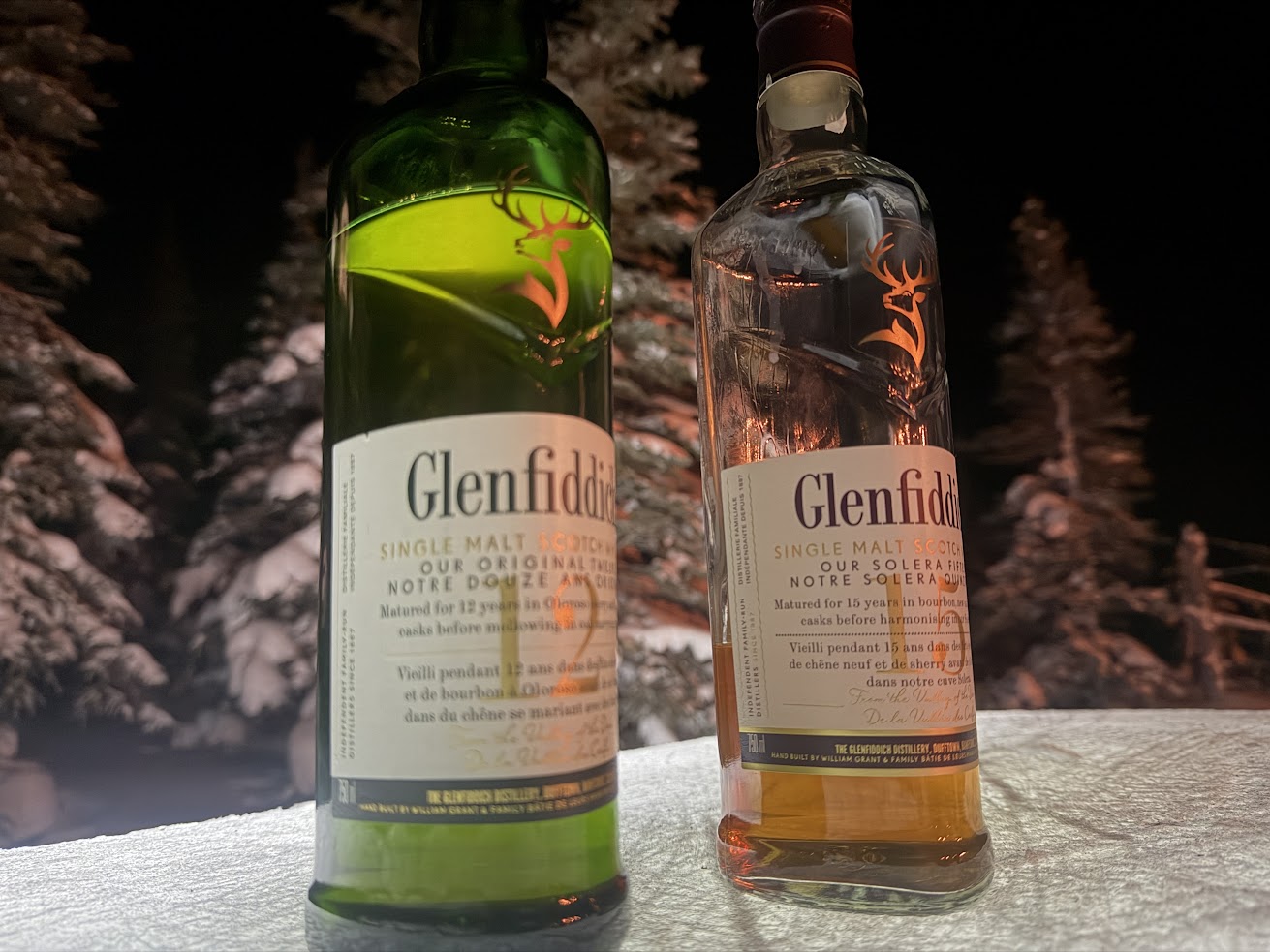 Two Glenfiddich whisky bottles on snow with snowy trees and a dark night background, evoking the magic of a Churchill Northern Lights tour.