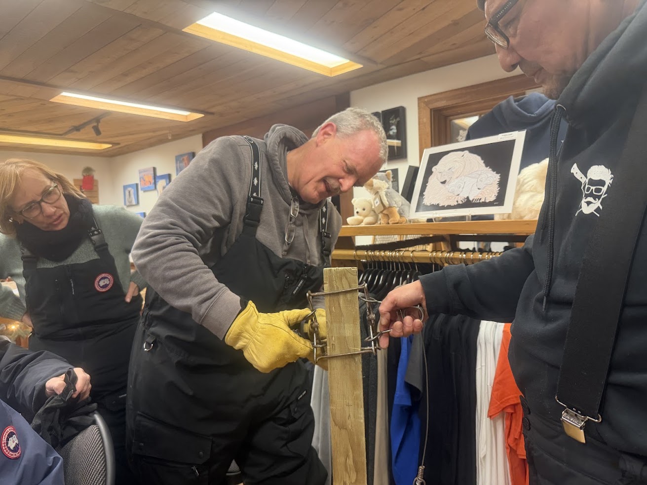 Three people in winter gear work together on a wooden post indoors, adjusting a metal mechanism&mdash;a behind-the-scenes glimpse into preparations for an unforgettable Churchill Northern Lights tour.