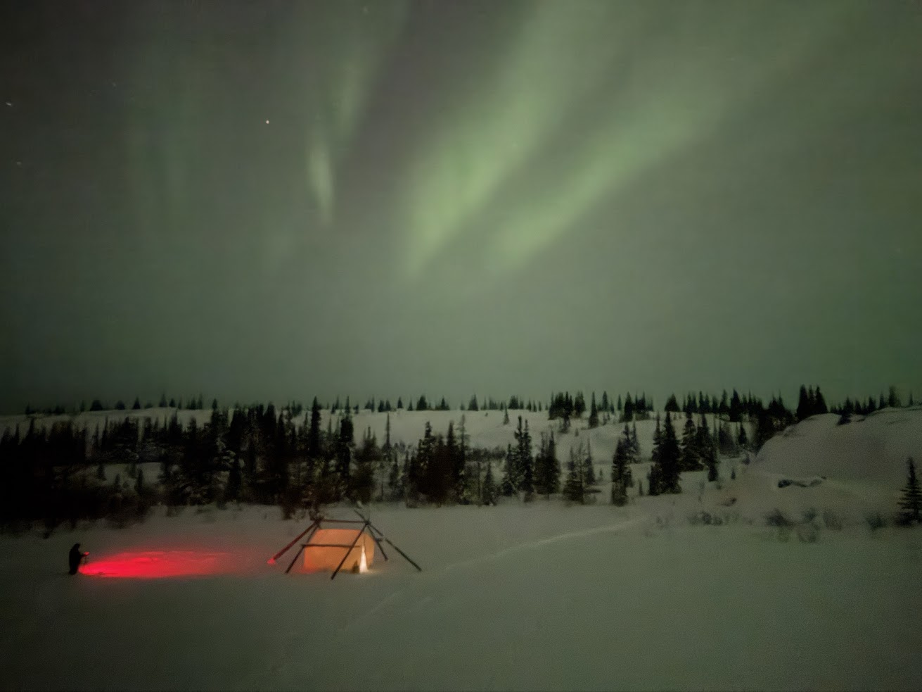 A snowy landscape under green northern lights, with a lit tent and a person holding a red light nearby&mdash;an enchanting scene reminiscent of a Churchill Northern Lights tour.