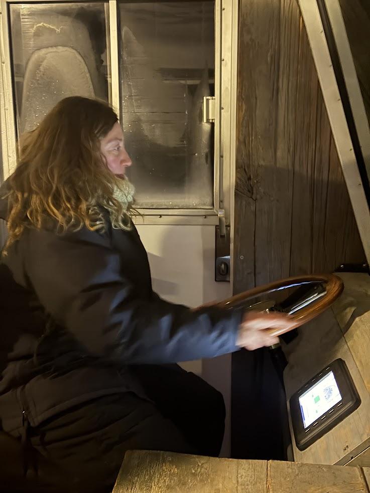 A person wearing a dark coat sits at a wooden steering wheel indoors, looking intently ahead, as if ready to embark on a Churchill Northern Lights tour.