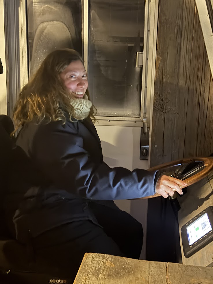 Woman in a coat and scarf smiling while sitting at the controls of a vehicle at night, ready to embark on an unforgettable Churchill Northern Lights tour.