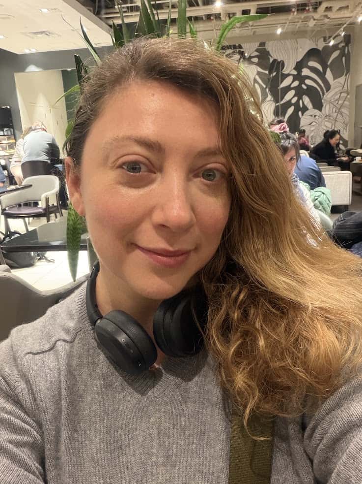Woman with wavy hair and headphones smiles in a caf&eacute;, browsing a Jackson Hole travel guide, with plants and people in the background.