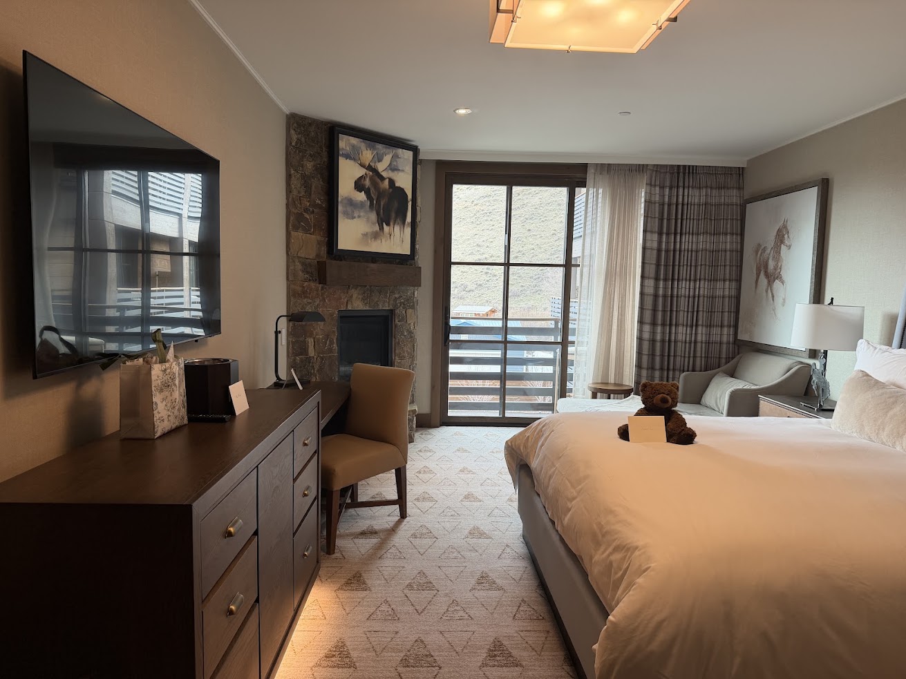 Modern hotel room at Rusty Parrot Lodge and Spa with a bed, TV, dresser, armchair, fireplace, large window, and elegant wall art.