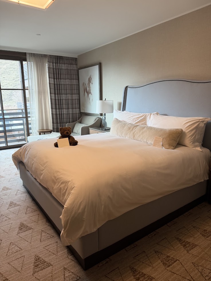 A neatly made hotel bed with a teddy bear and note, near a window and armchair, exudes the warm hospitality of the Rusty Parrot Lodge and Spa.
