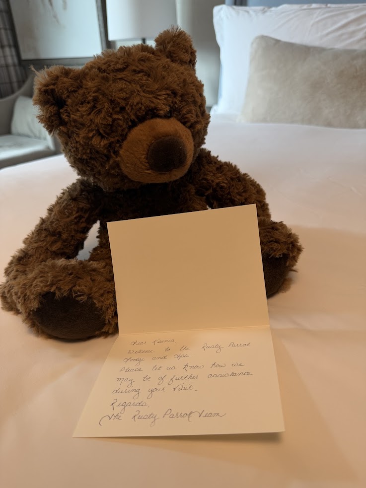 A brown teddy bear sits on a bed behind an open handwritten card with a message inside, creating a cozy welcome reminiscent of the thoughtful touches found at the Rusty Parrot Lodge and Spa.
