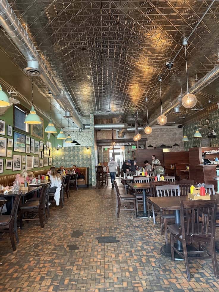 Restaurant interior with wooden tables, vintage lights, people dining, and a patterned tile floor&mdash;just the kind of cozy spot you'll find recommended in any great Jackson Hole travel guide.