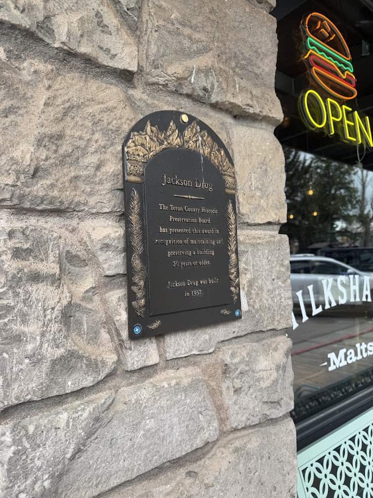 A historic plaque on a stone wall near a window with a neon "OPEN" sign and reflections of street trees&mdash;an inviting scene often highlighted in any comprehensive Jackson Hole travel guide.
